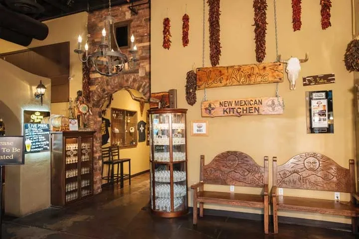 A restaurant lobby with wooden benches, Southwestern decor, hanging dried peppers, and signs reading “New Mexican Kitchen” and “Please Wait to Be Seated”—a warm space common in commercial electrical projects Arizona.
