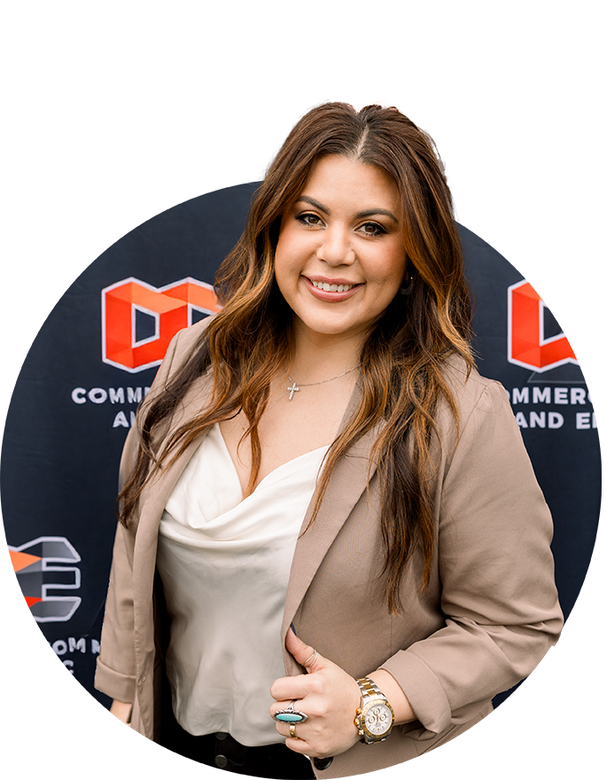 A woman with long brown hair wearing a beige blazer and white blouse stands smiling in front of a Commercial Comm and Electric backdrop with orange and white geometric logos.