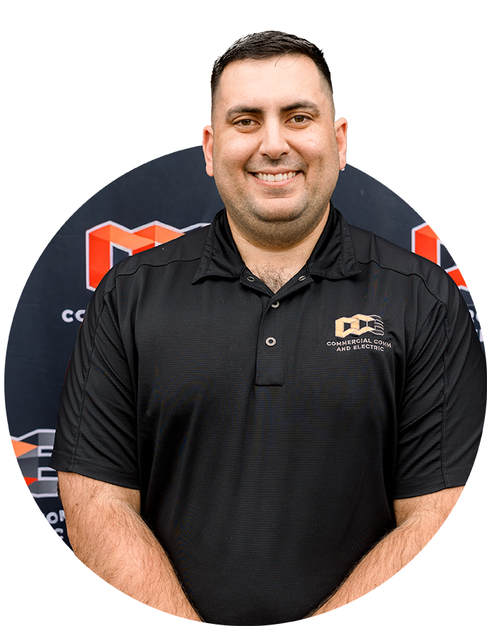 A man in a black polo shirt with the Commercial Comm and Electric logo stands in front of a branded backdrop, smiling at the camera.